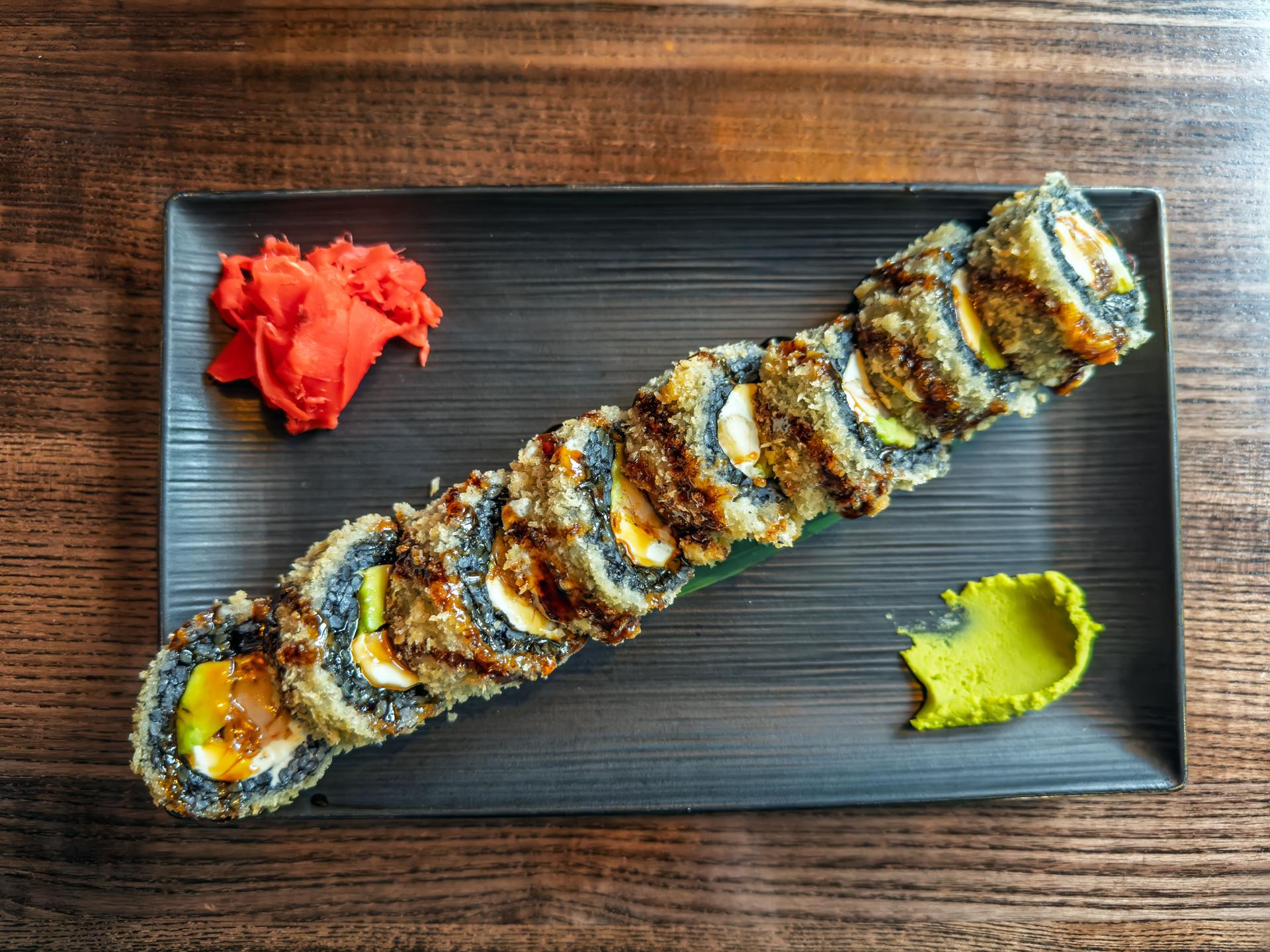 Tempura Sushi Roll with Ginger and Wasabi on Black Plate Served on Wooden Table.Crispy Fried Sushi Roll with Traditional Condiments on Dark Rectangular Plate, top view